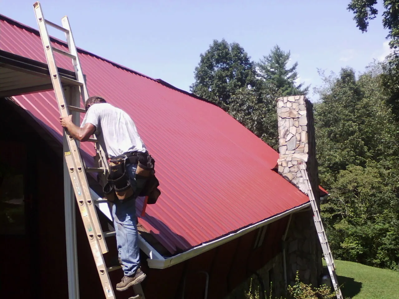 Roofer on a ladder beside a red metal roof and stone chimney — Soffit & Fascia Repair by Roofing Brothers in Takoma Park
